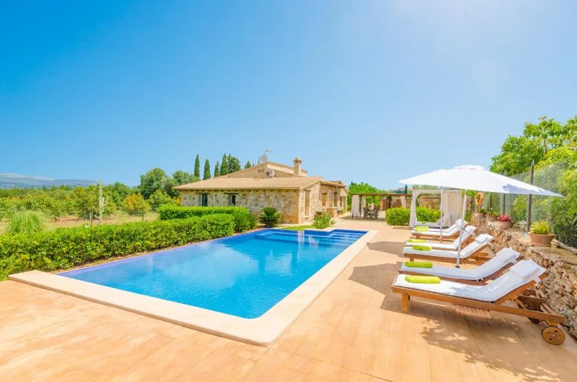 An outdoor pool beside a stone house, with sun loungers under an umbrella.