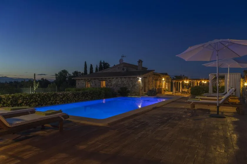 A lit swimming pool beside a wooden deck, lounge seating, and patio umbrellas at dusk.