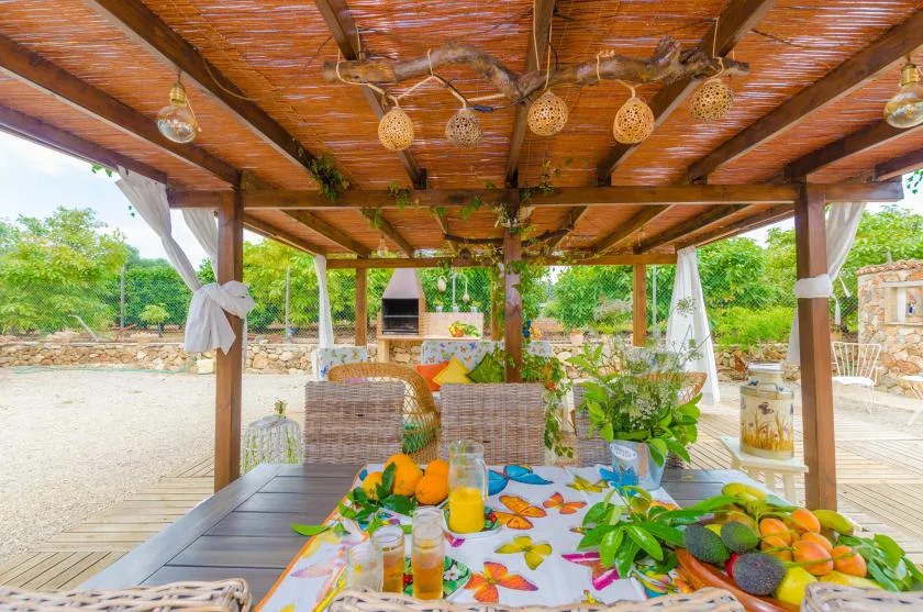 A wooden pergola shelters a dining table set with fruit and drinks in a garden.