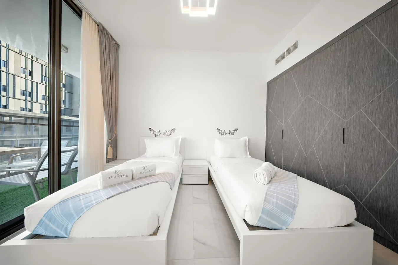 A bright bedroom with two beds beside floor-to-ceiling windows.