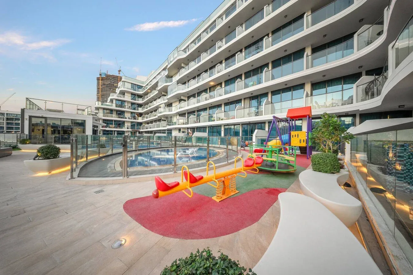 A courtyard playground with colorful play equipment sits beside a glass-railed pool.