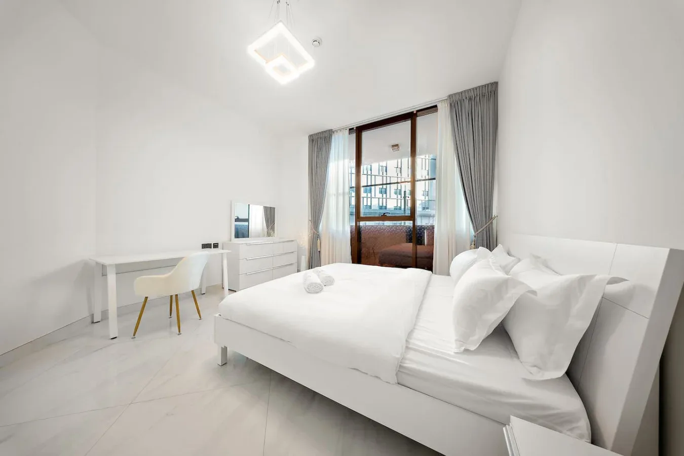 A bright bedroom with a white bed, a desk, and a glass door.