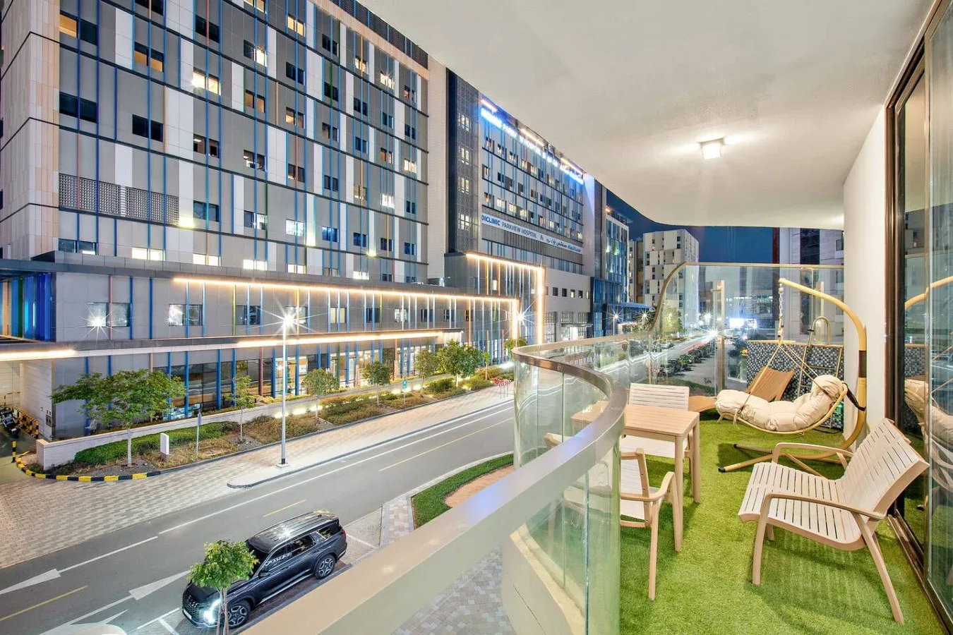 A furnished balcony with green flooring overlooks a street and modern buildings.