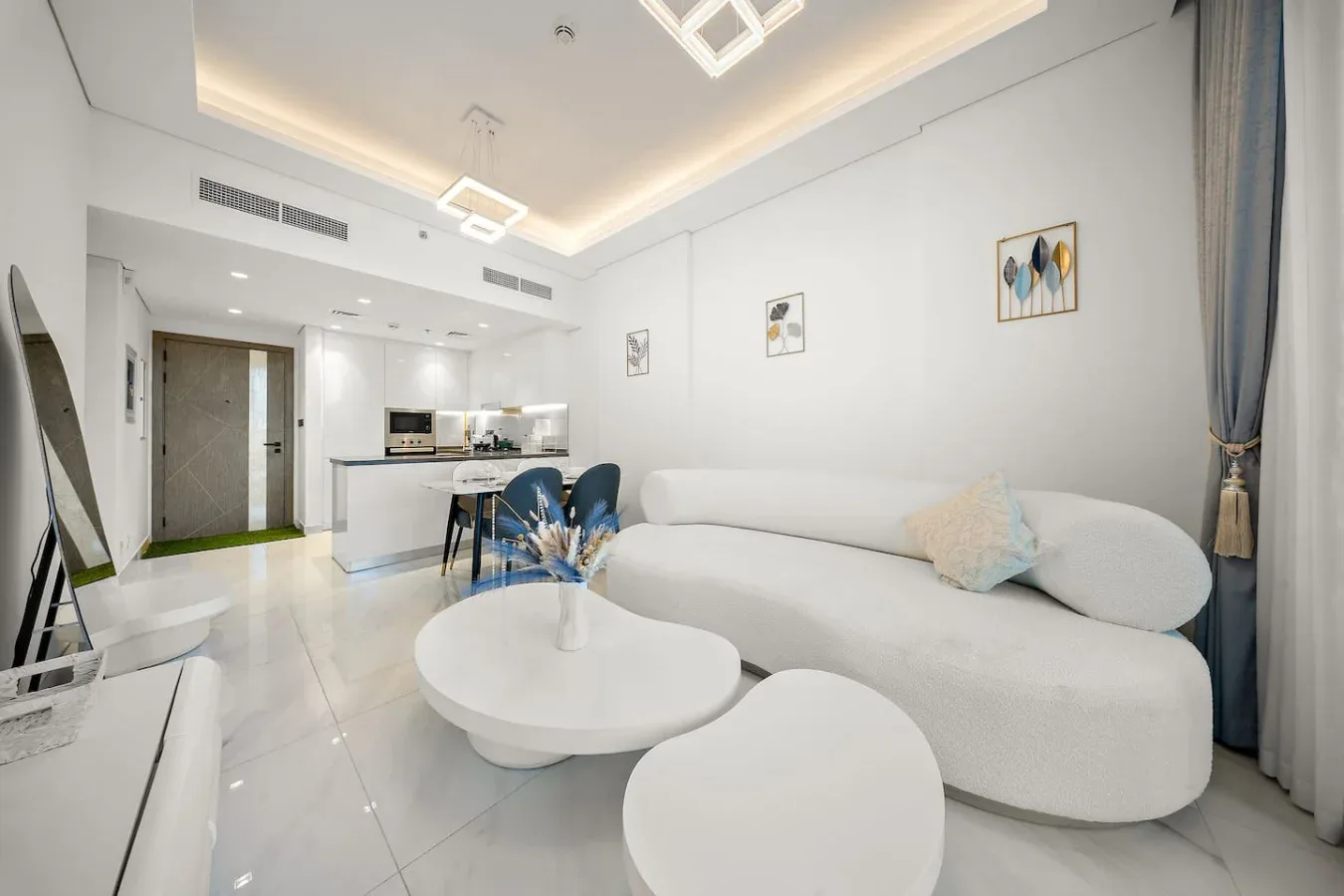 A bright living room with white seating, round tables, and a compact kitchen area.