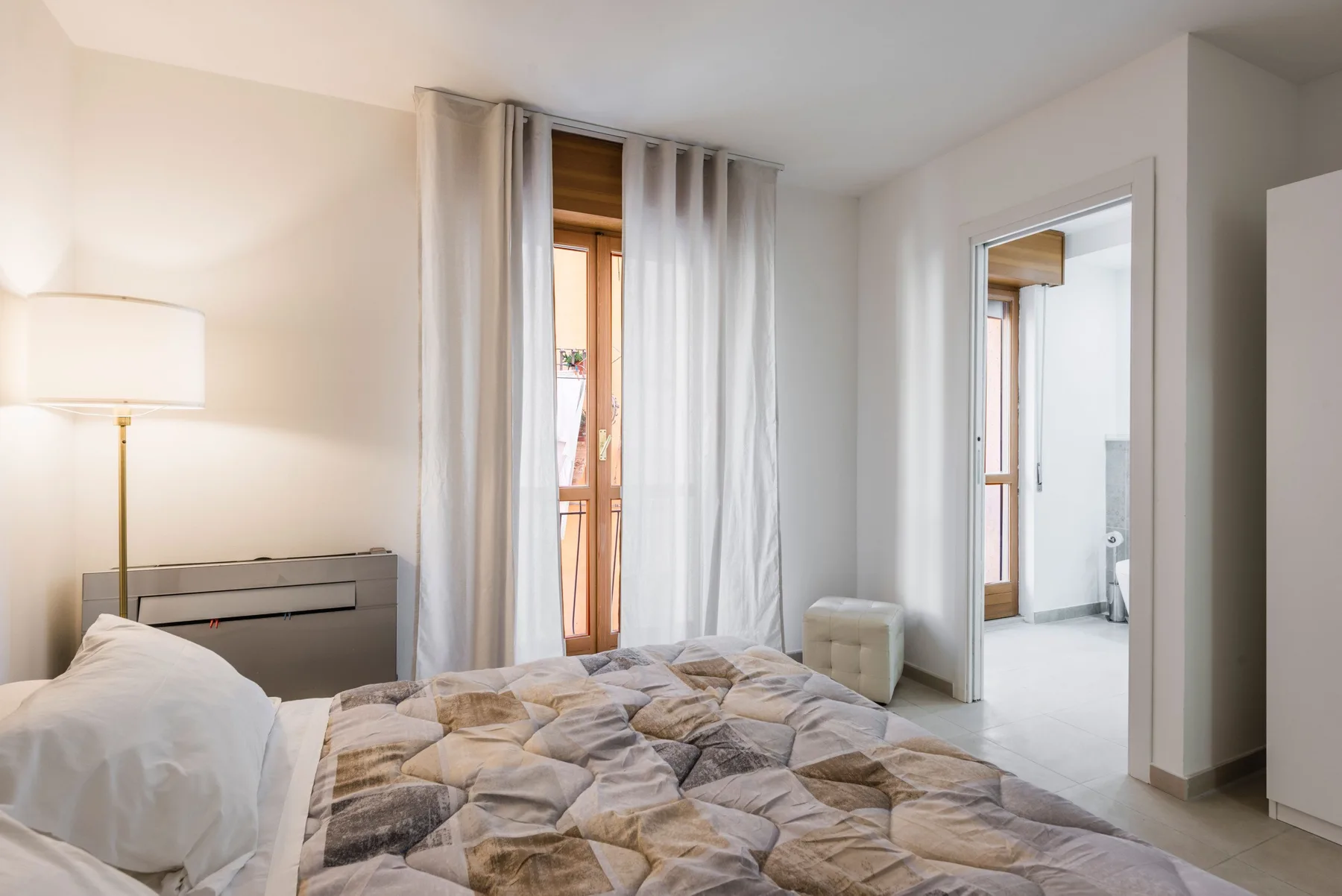 Un dormitorio con la cama deshecha, cortinas y una puerta abierta hacia el baño.