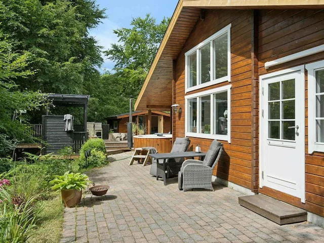 A wooden house opens onto a paved patio with outdoor seating and surrounding greenery.