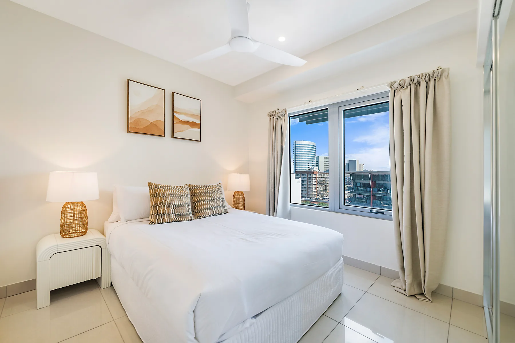 A bright bedroom has a neatly made bed beside a window overlooking city buildings.