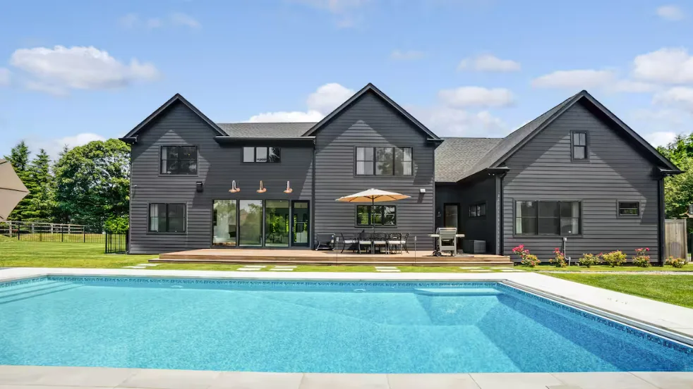 A large outdoor swimming pool sits beside a modern dark-sided house.