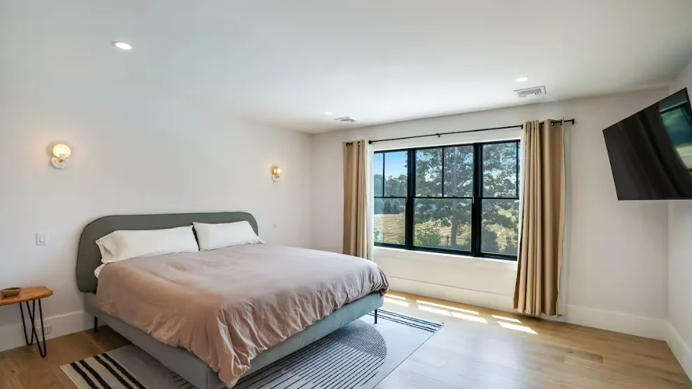 Dormitorio con una cama junto a una ventana grande y un televisor montado en la pared.