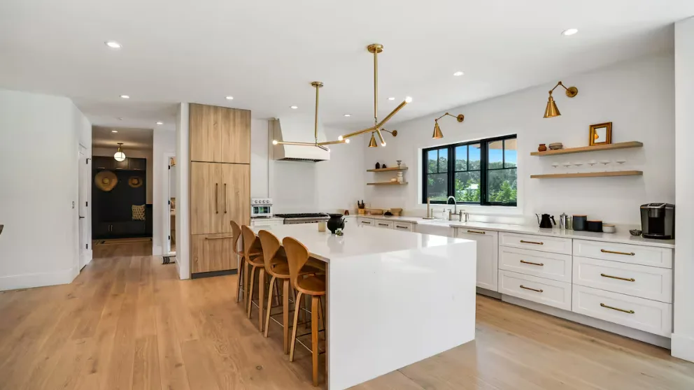 Una cocina moderna y luminosa con una isla blanca, asientos de madera y ventanas grandes.