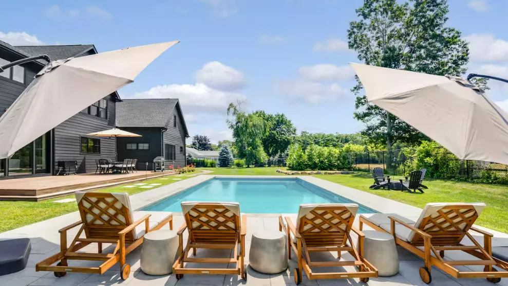 Una piscina al aire libre junto a una casa moderna, con asientos de descanso junto al borde.