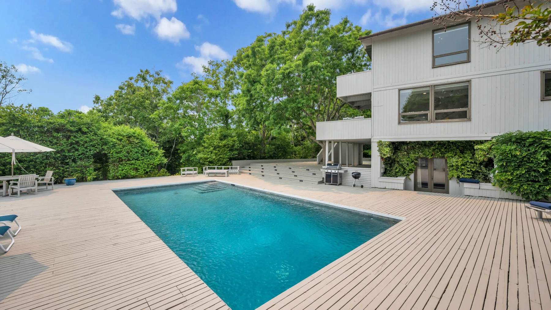 Una piscina al aire libre está junto a una casa blanca, rodeada por una amplia terraza de madera.