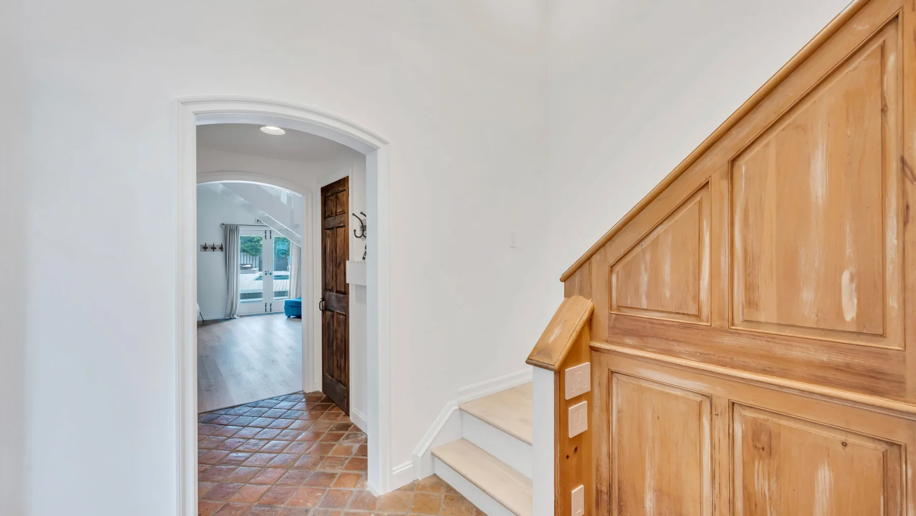 Un pasillo con suelo de baldosas conduce a una habitación luminosa junto a una escalera de madera.
