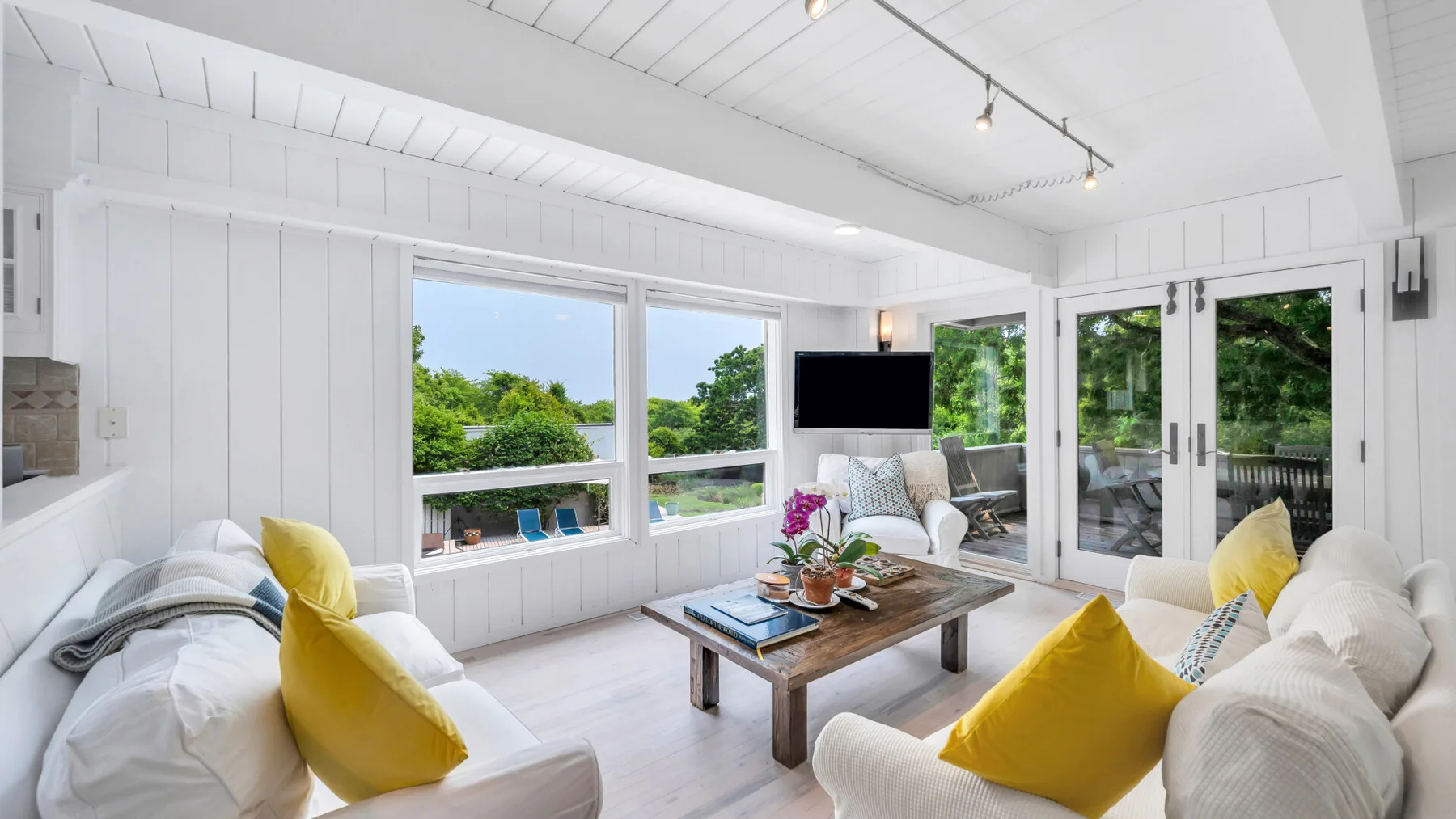Una sala luminosa tiene asientos blancos, cojines amarillos y grandes ventanas con vistas a la vegetación.