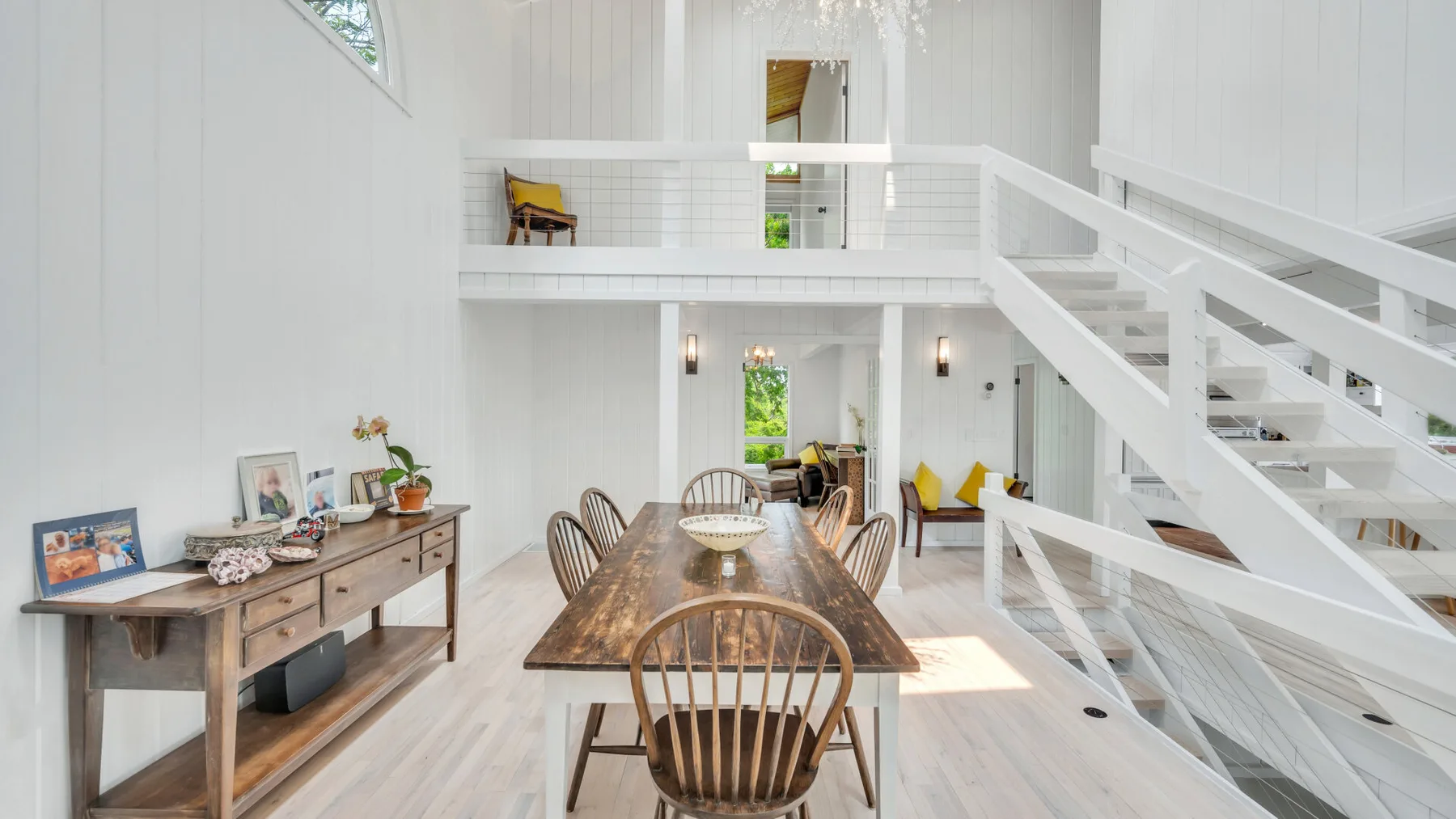 Un comedor luminoso con una mesa de madera bajo una escalera blanca y un rellano superior.