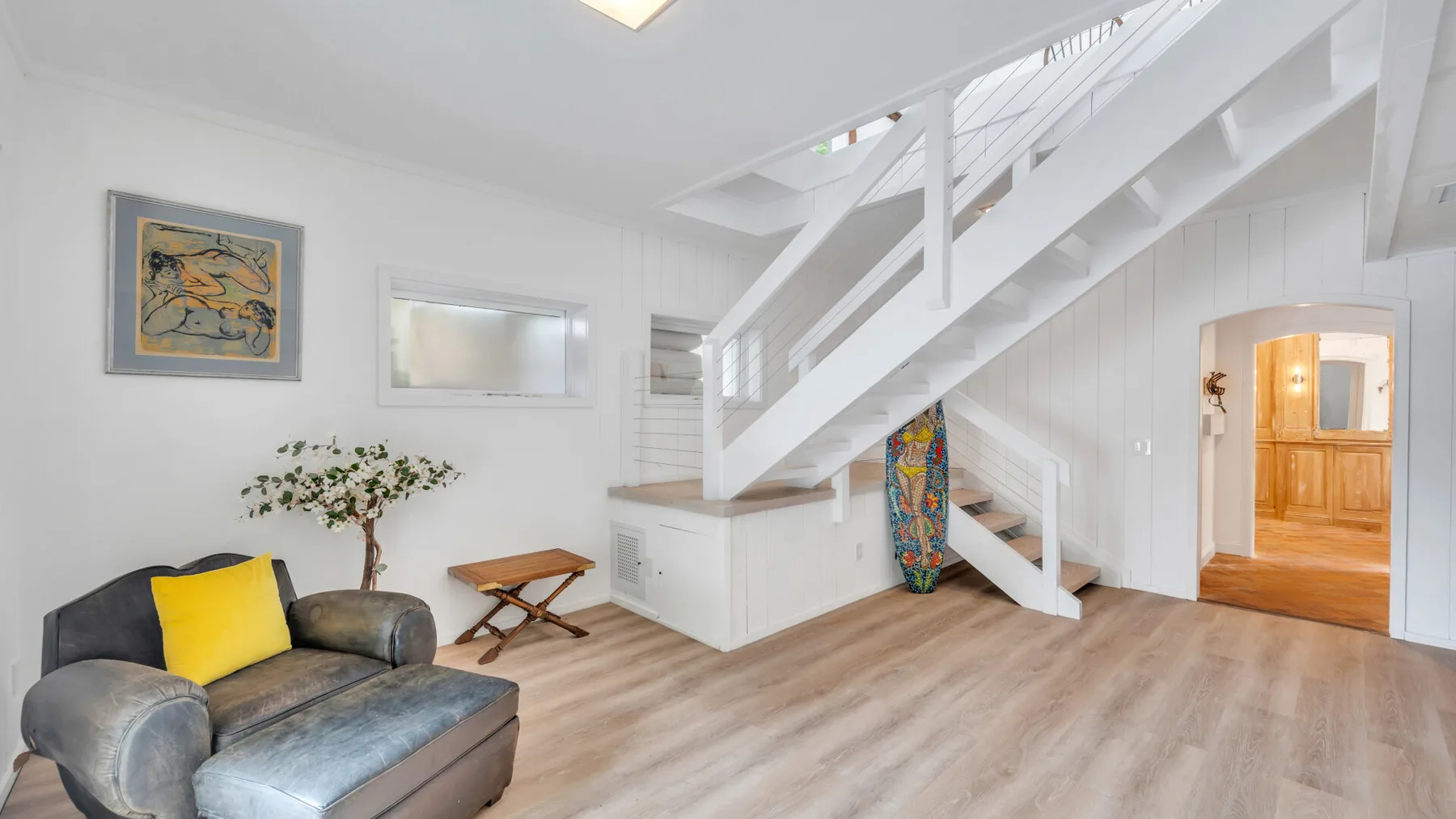 Una sala luminosa con asientos bajo una escalera blanca y una puerta abierta.