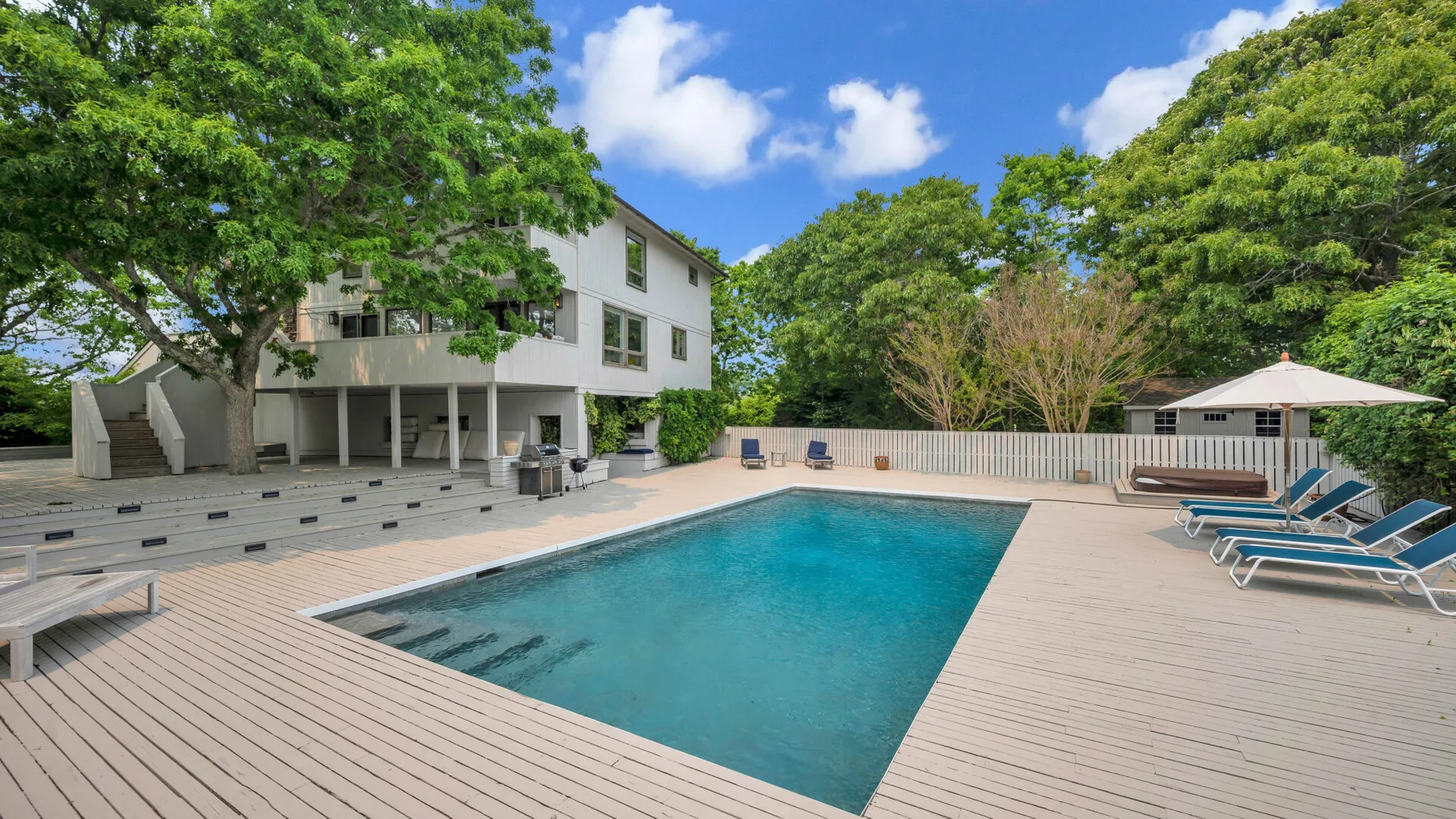 Una piscina al aire libre está junto a una casa moderna, rodeada de árboles y asientos de descanso.