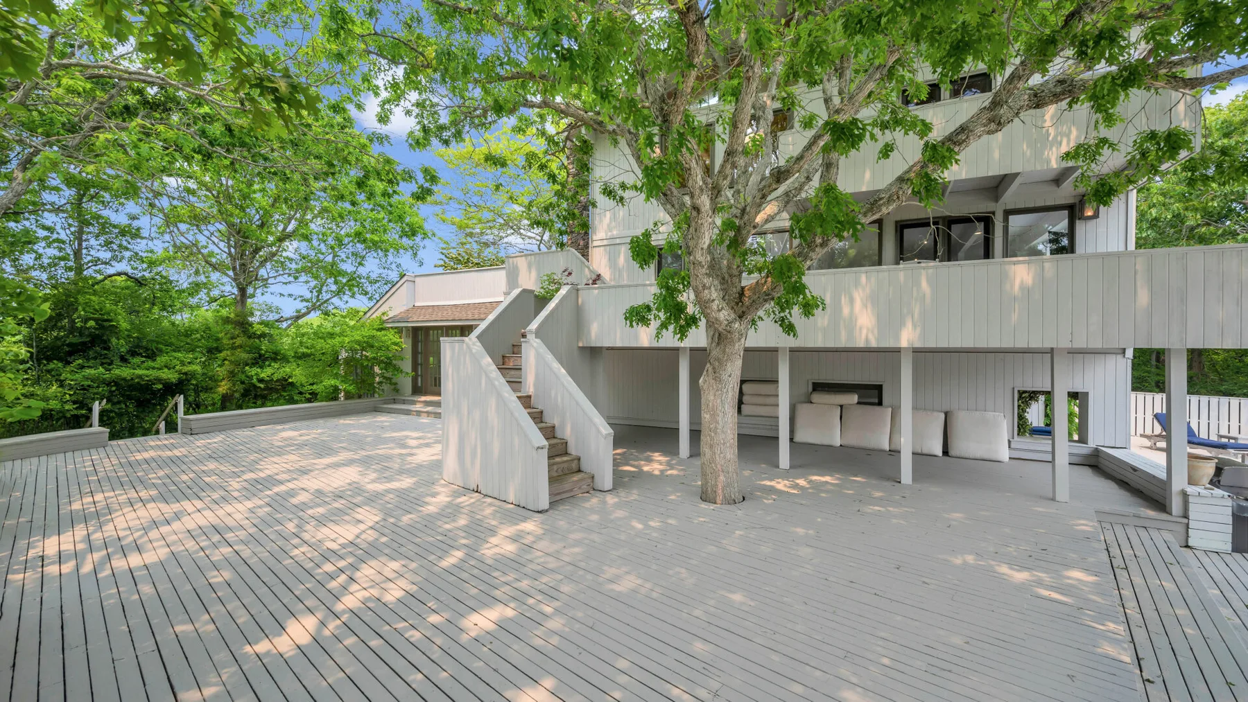 Una casa moderna se abre a una amplia terraza de madera, sombreada por árboles frondosos.