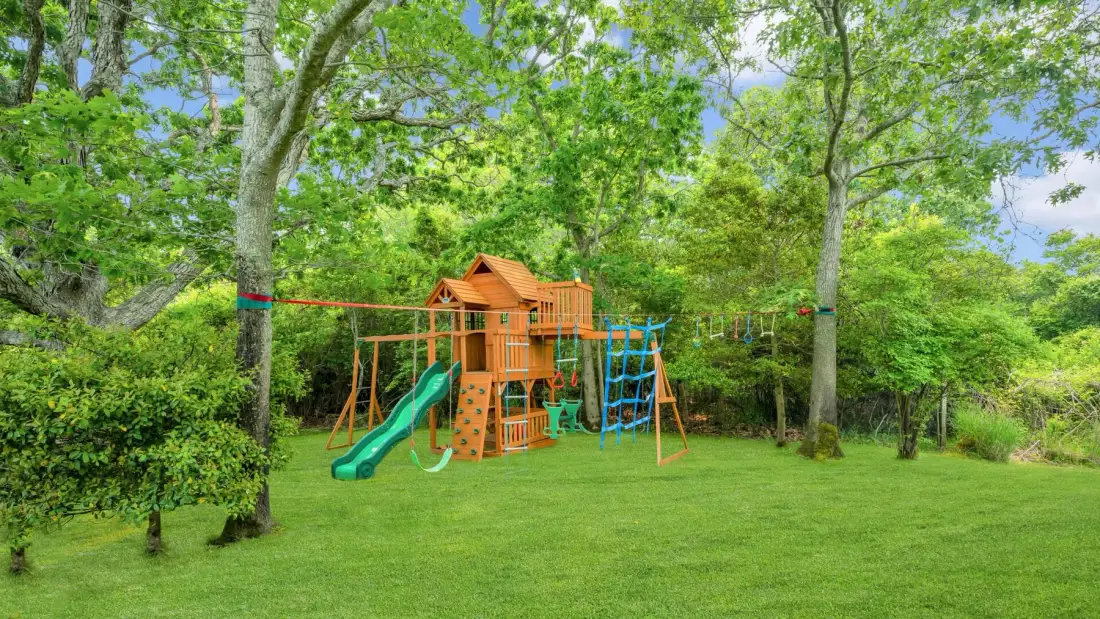 Un parque infantil de madera con toboganes y estructuras para trepar está en un césped rodeado de árboles frondosos.