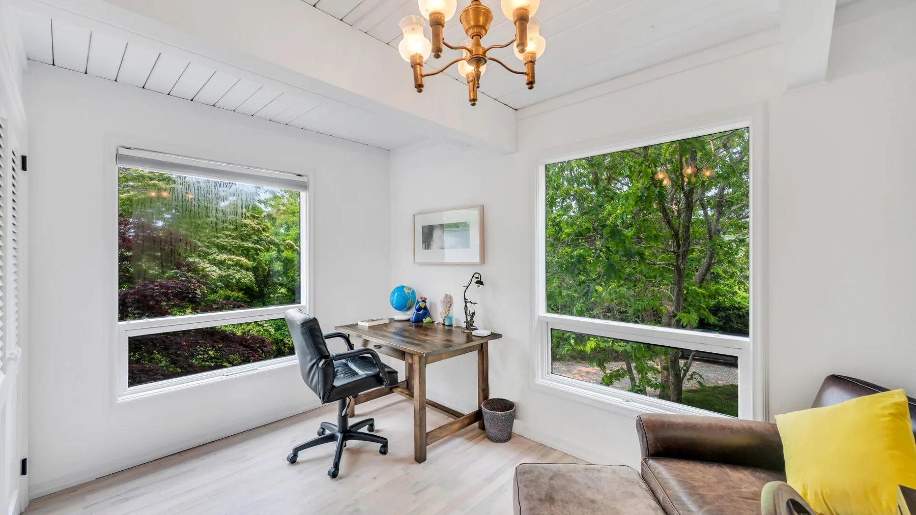 Un estudio luminoso con escritorio y silla junto a grandes ventanas con vistas a la vegetación.