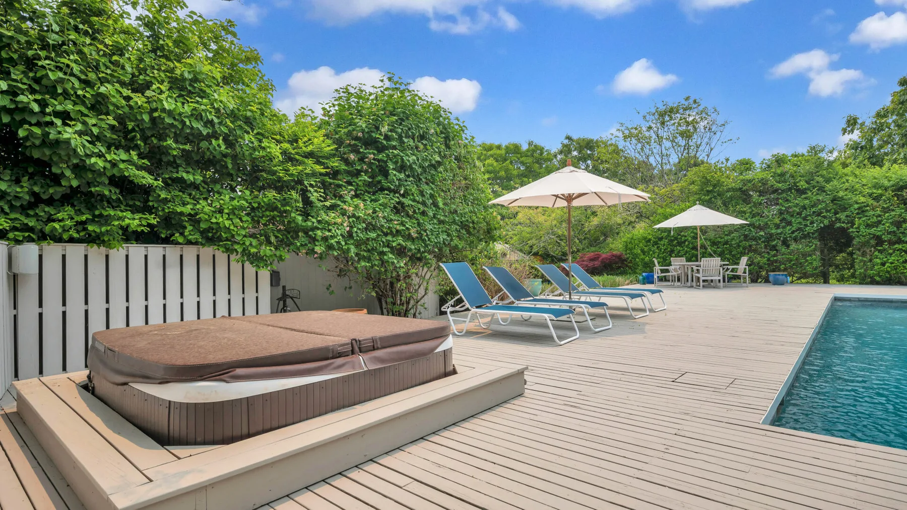 Una terraza junto a la piscina tiene asientos reclinables bajo sombrillas, rodeada de vegetación.