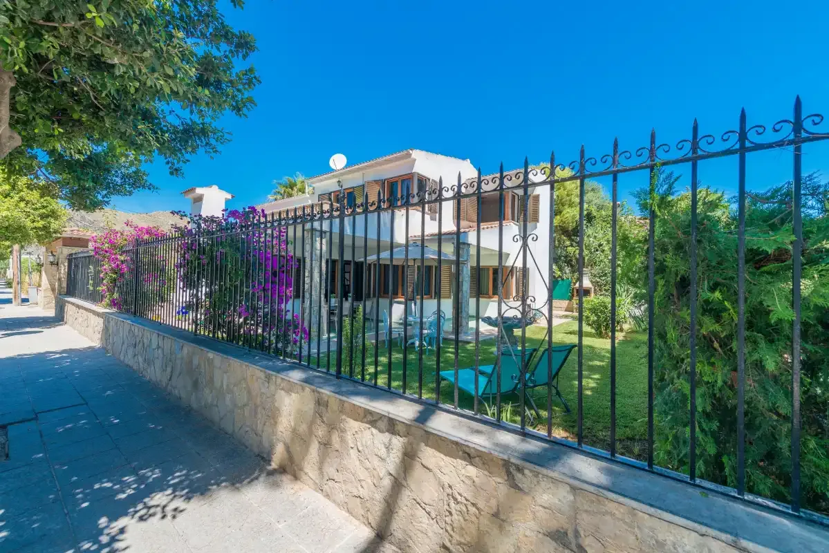 Una casa moderna y un jardín se ven tras una verja metálica negra junto a una acera.
