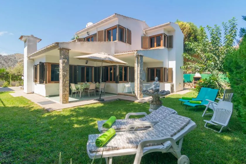 Una casa de dos plantas da a un jardín con césped, asientos y tumbonas.