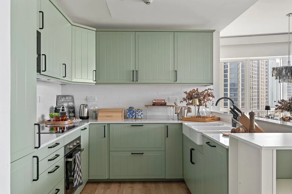 A modern kitchen with pale green cabinets, white countertops, and a large window.