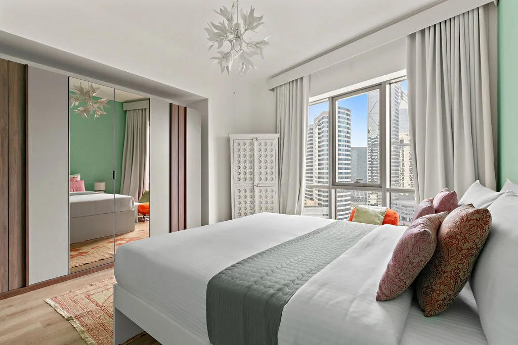 A bright bedroom with a large bed beside windows overlooking city buildings.