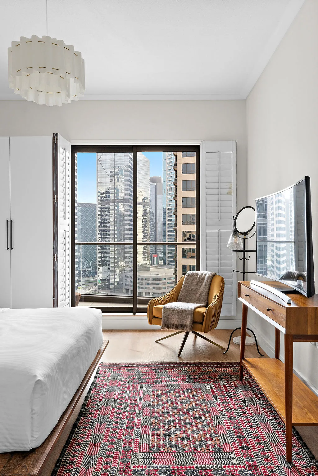 A bedroom with a bed, armchair, and large windows overlooking city buildings.