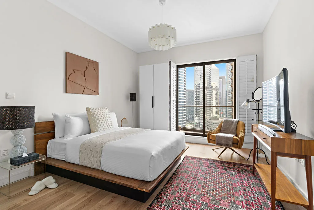 A bright bedroom has a large bed, patterned rug, and floor-to-ceiling windows.
