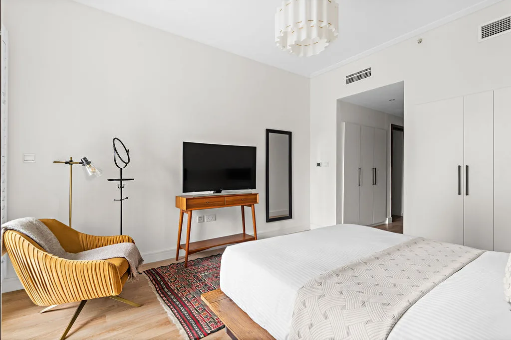 A bright bedroom features a bed, a television on a wooden stand, and an upholstered seating area.