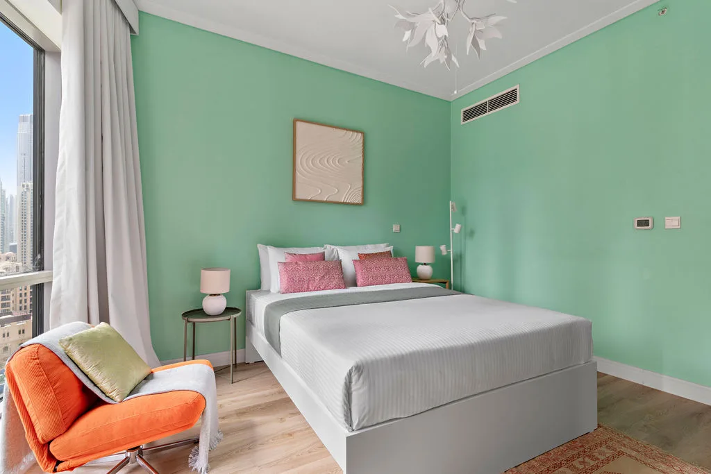 A bedroom with a bed, mint-green walls, and an orange chair beside the window.