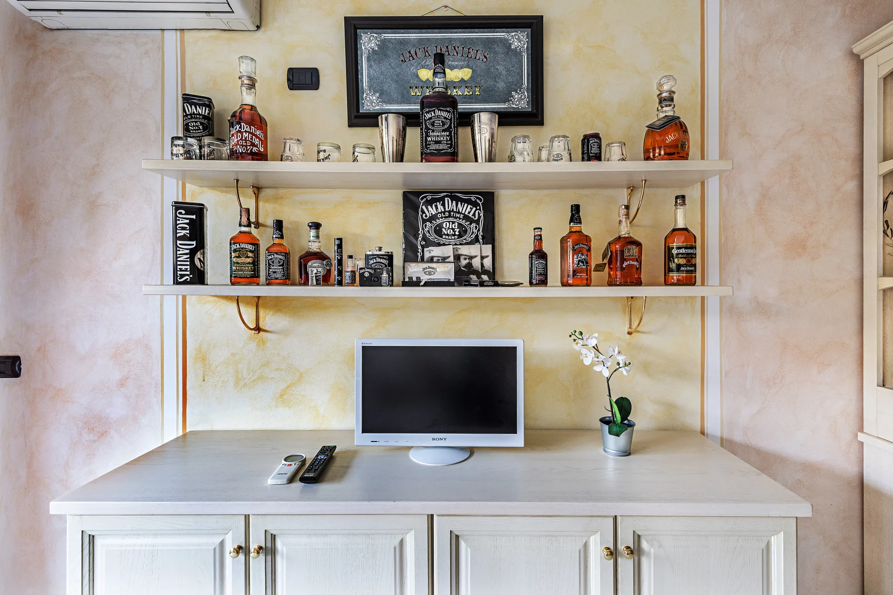 Un mueble con estantes exhibe botellas sobre una encimera con un monitor y una planta.