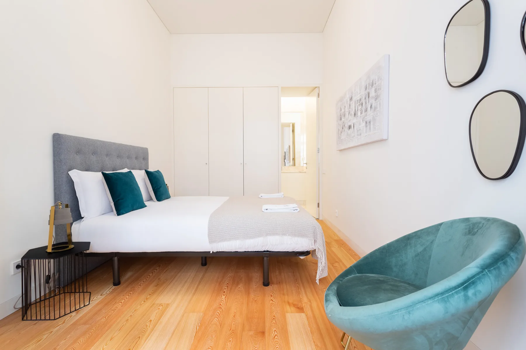 Dormitorio luminoso con una cama, cojines verde azulado y un sillón redondeado.