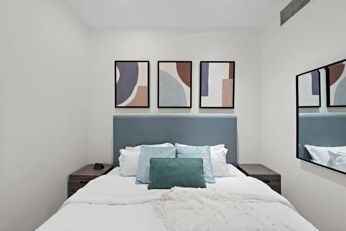 A bedroom with a neatly made bed, colorful abstract artwork, and a wall mirror.
