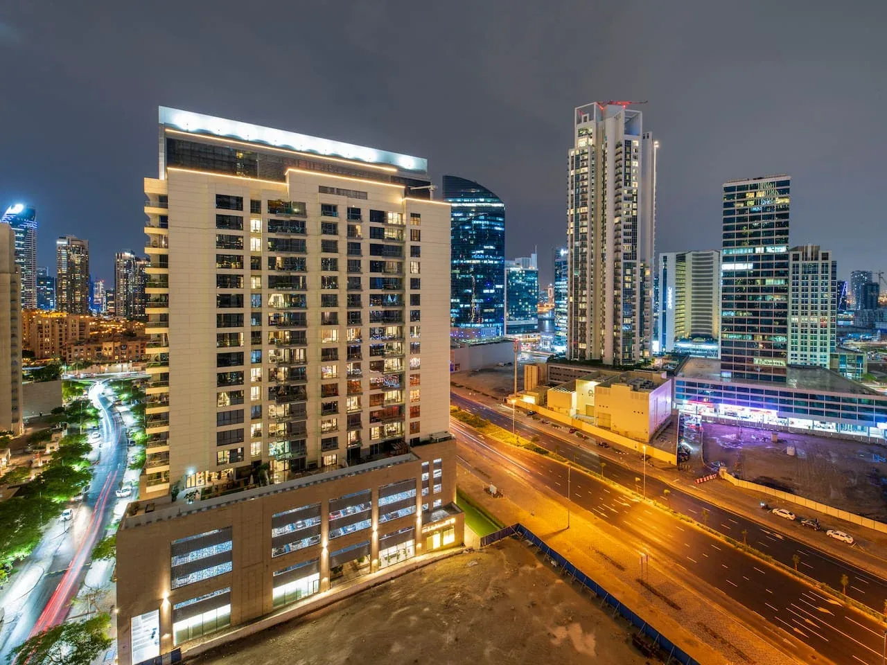 A nighttime cityscape shows illuminated high-rise buildings beside a busy road.