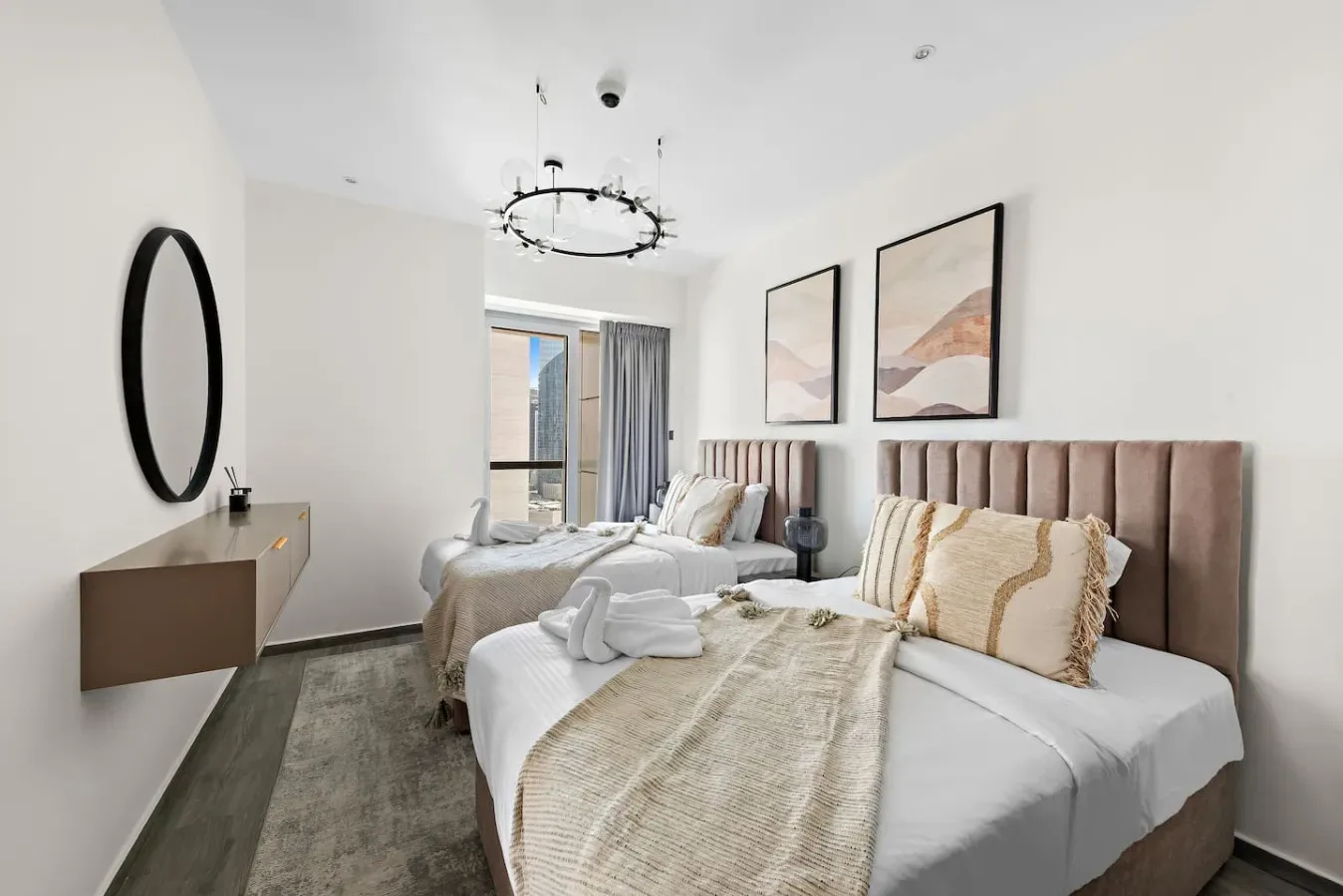 A bright bedroom with two beds, neutral bedding, and framed wall art.