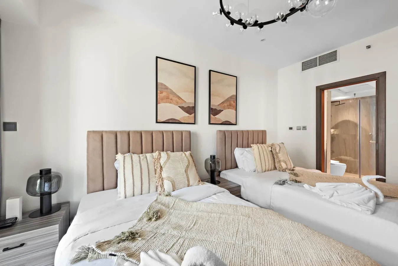 A bedroom with two beds, neutral bedding, and framed artwork above them.