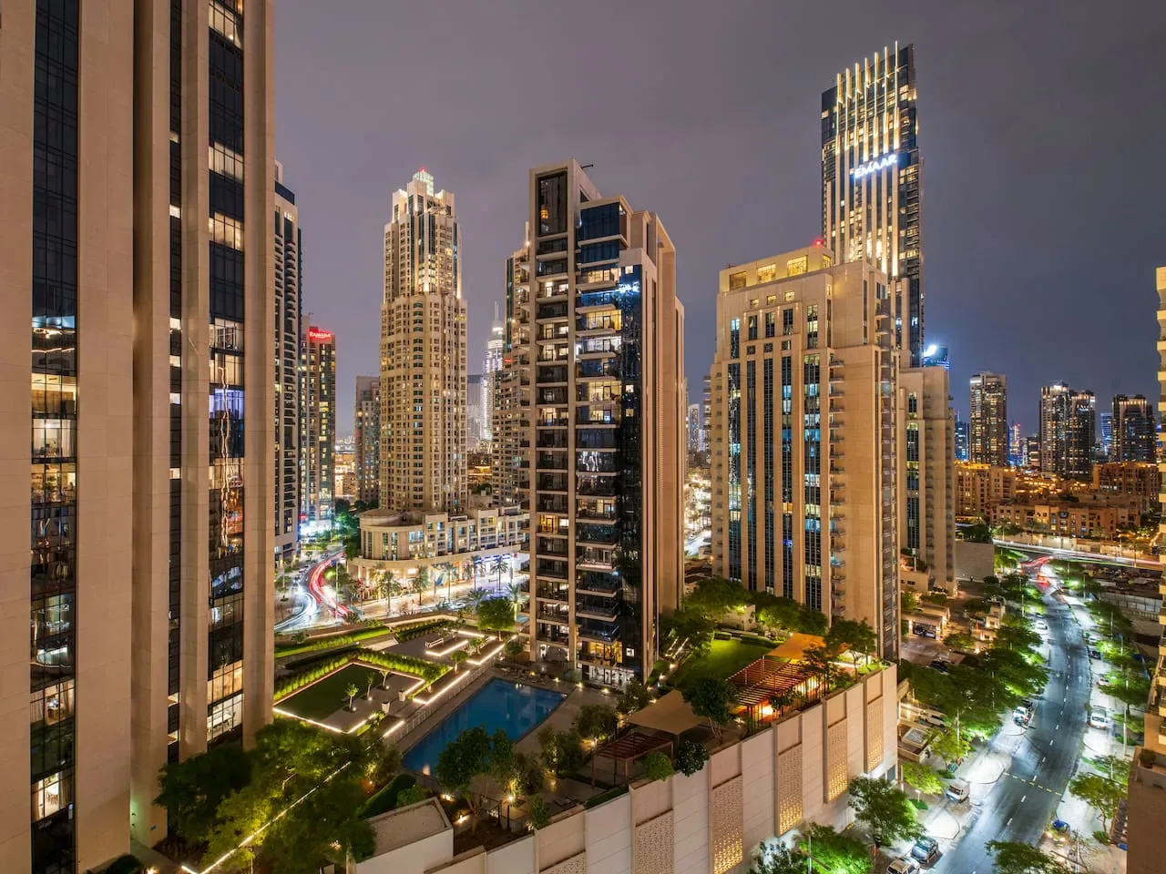A nighttime city skyline of illuminated high-rise buildings surrounds a landscaped courtyard.