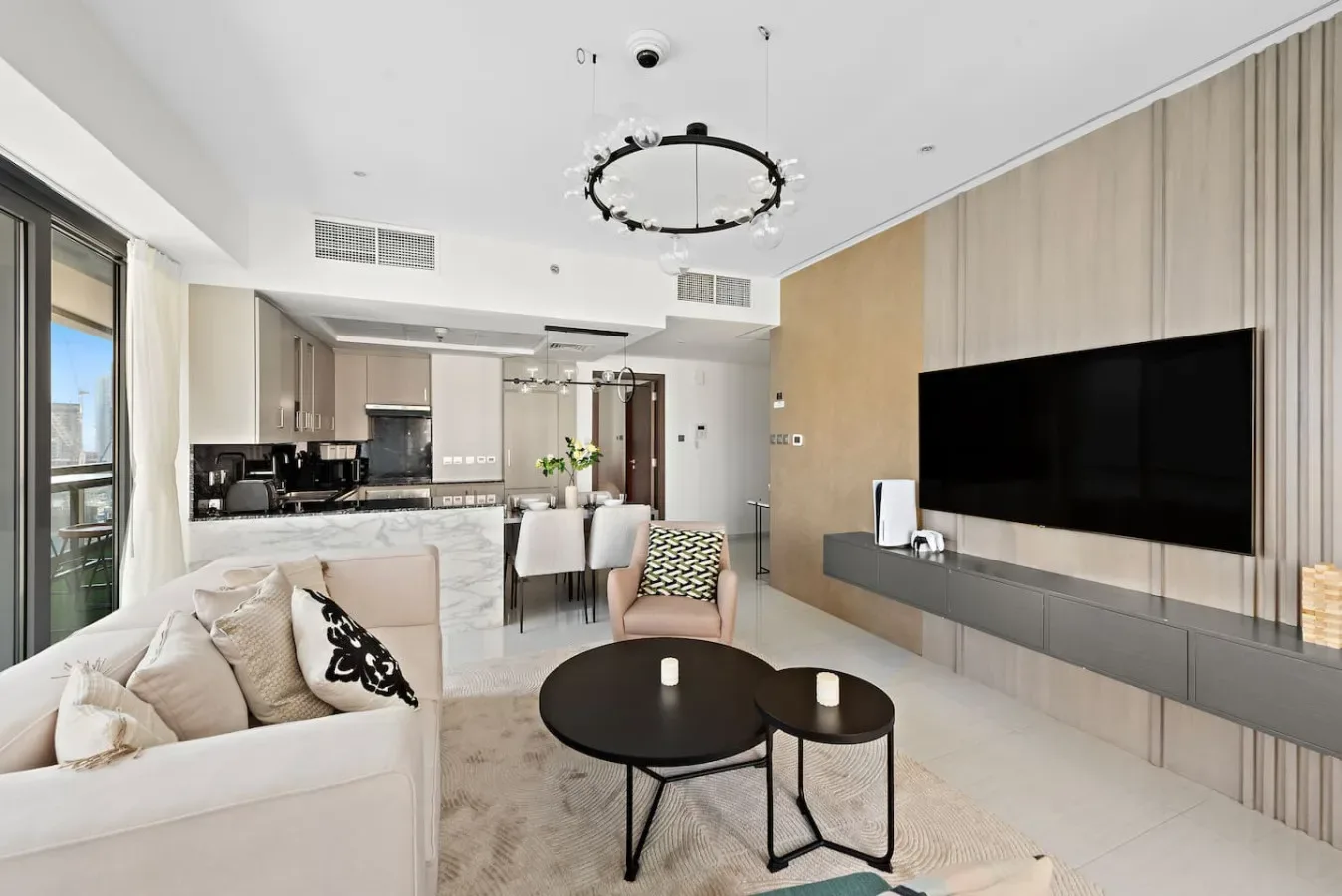 A bright living room opens onto a dining area and kitchen, with a large wall-mounted television.