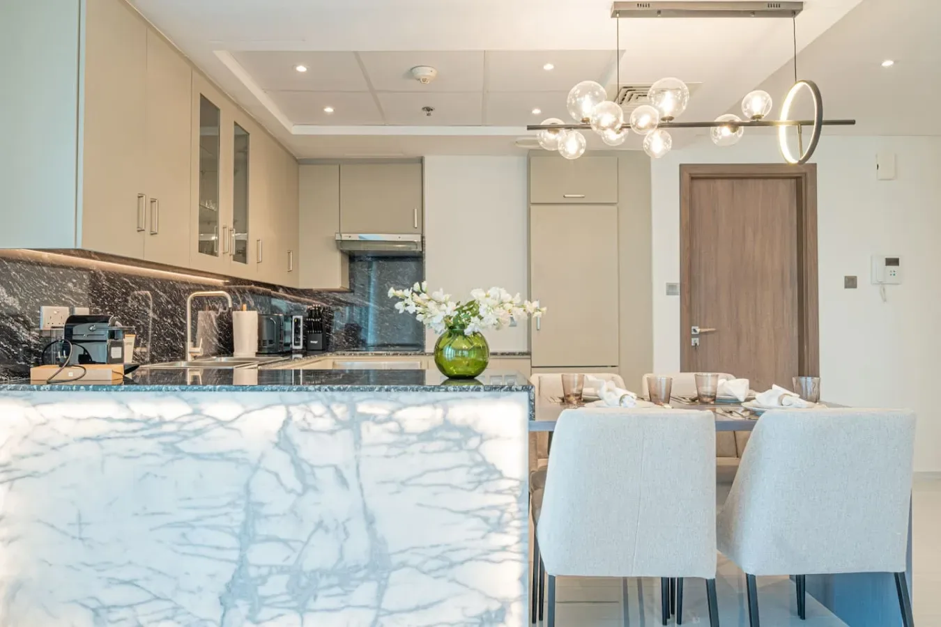 A modern kitchen and dining area with pale cabinetry, a marble-patterned island, and dining chairs.