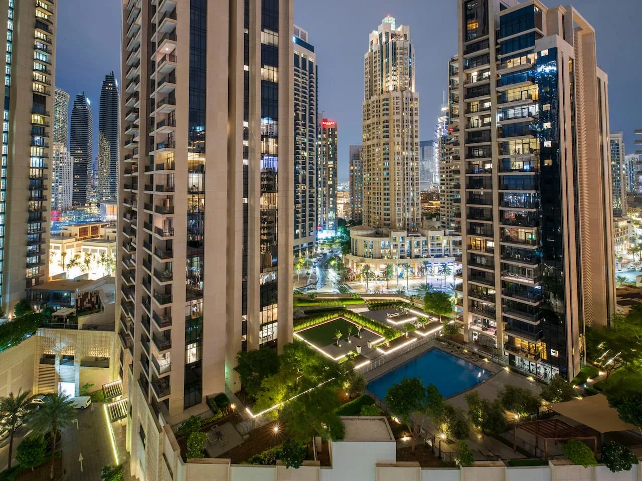 A nighttime cityscape of illuminated high-rise buildings surrounding a landscaped courtyard and pool.