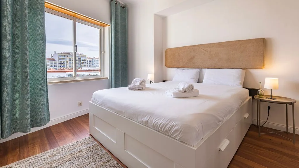 Un dormitorio con una cama grande junto a una ventana con vistas a edificios cercanos.
