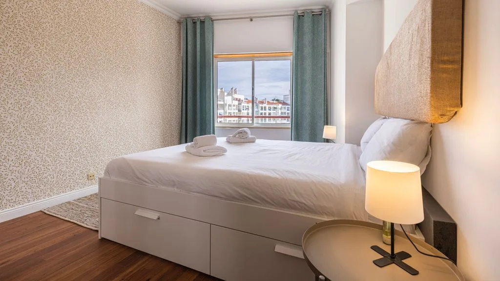 Un dormitorio con cama, mesita de noche y una ventana con vistas a edificios cercanos.