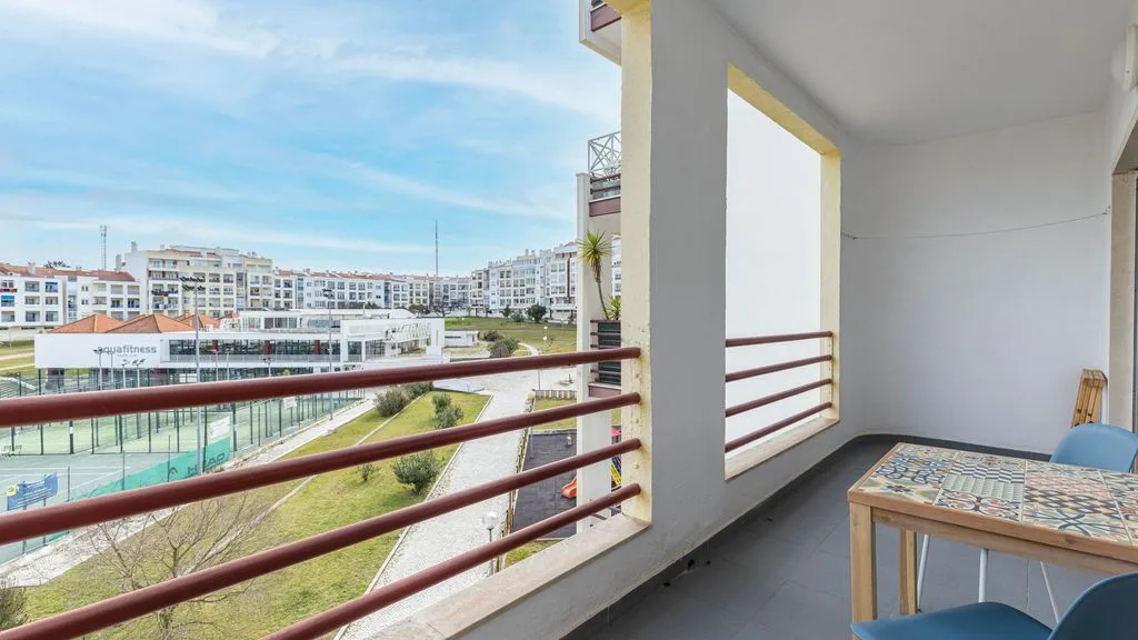 Un balcón cubierto tiene una mesa y sillas con vistas a edificios cercanos y una cancha deportiva.