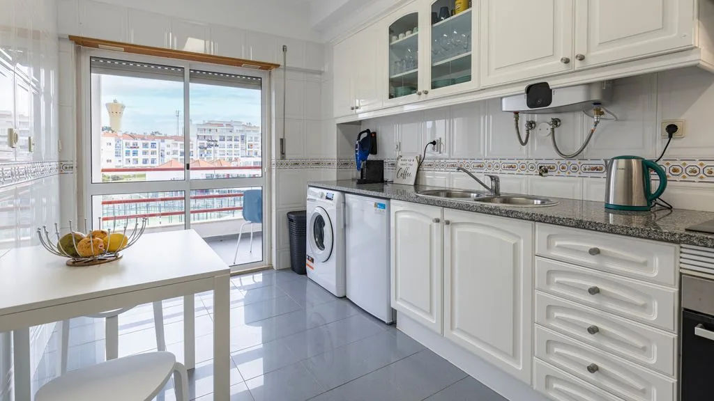 Una cocina luminosa tiene armarios blancos, una encimera y una mesa junto a una ventana grande.