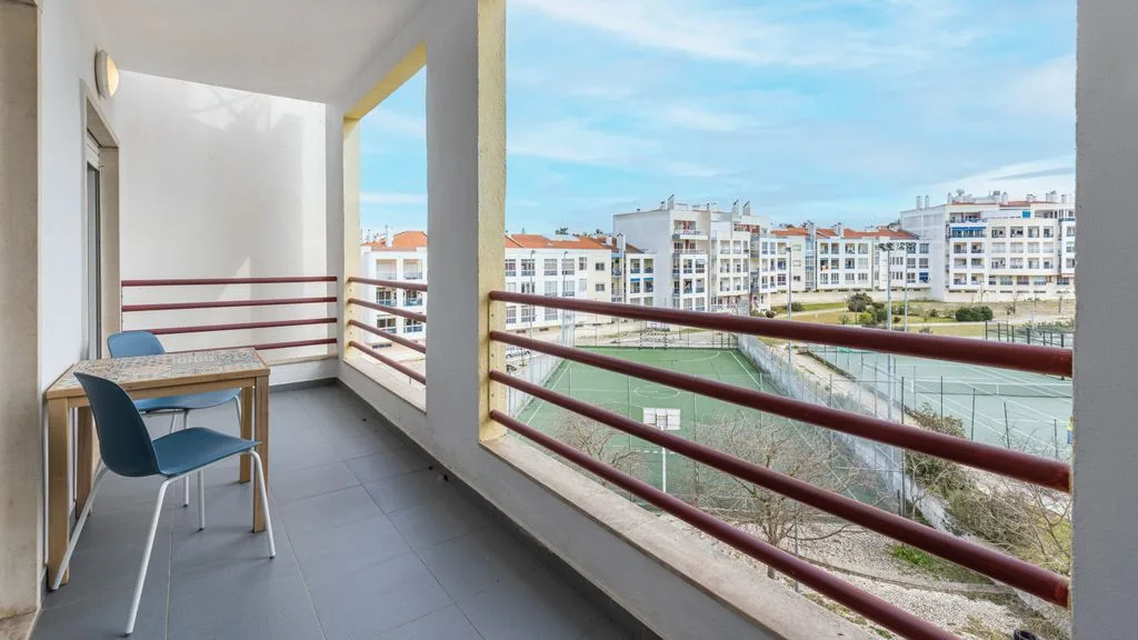 Un balcón cubierto con una mesa pequeña y sillas tiene vistas a edificios cercanos.