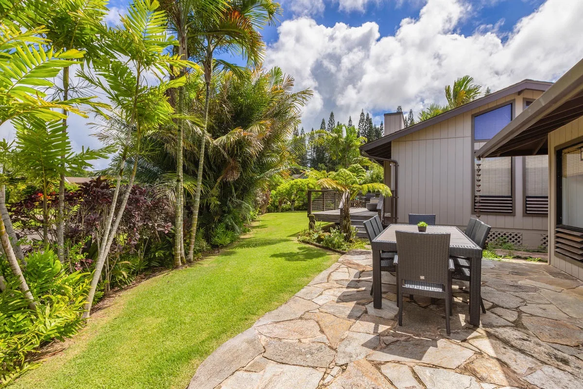 A stone patio with outdoor seating sits beside a green lawn and tropical plants.