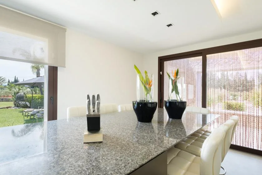 A bright dining area with a long table, cushioned seating, and potted plants.